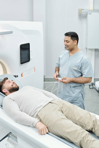 Szczęśliwy mieszany lekarz w mundurze medycznym i uśmiechnięty brodaty pacjent patrzący na siebie po badaniu w ultradźwiękowej maszynie - Zdjęcie, obraz