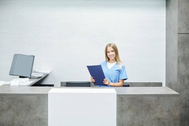 Feliz joven asistente bonita o recepcionista en uniforme azul sujetando portapapeles con documento médico mientras está de pie junto al mostrador en las clínicas - Foto, imagen