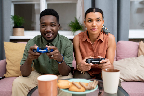 Menino adolescente feliz de etnia africana e menina mestiça sentada no sofá contra a janela na sala de estar e jogando videogame - Foto, Imagem