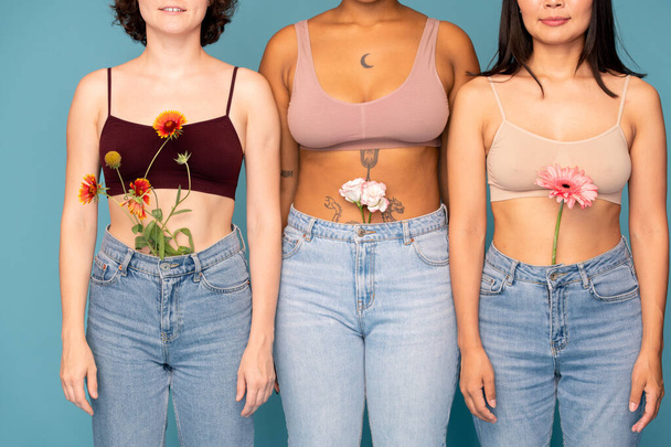 Front view of three young intercultural women with fresh flowers in jeans standing in row close to one another against blue background - Photo, Image