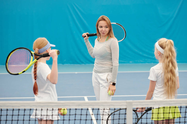 Νεαρή γυναίκα εκπαιδεύτρια τένις με αυτοπεποίθηση, που κρατάει ρακέτα πίσω από το λαιμό και κοιτάζει δύο κορίτσια ενώ τις συμβουλεύεται στο στάδιο. - Φωτογραφία, εικόνα