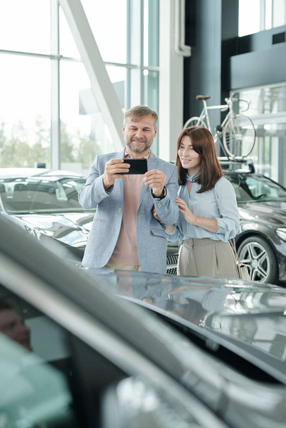 Glücklicher reifer Mann mit Smartphone, der ein Foto vom luxuriösen neuen Auto macht, während er neben seiner Frau vor dem Hintergrund der Auto-Center-Umgebung steht - Foto, Bild