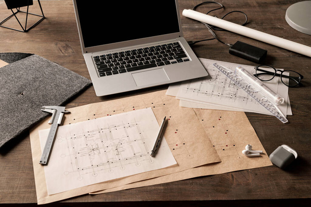 Laptop, narzędzia ręczne, ołówek, okulary i inne artykuły zaopatrzeniowe otoczone rozłożonymi planami z szkicami nowego budynku narysowanymi przez inżyniera - Zdjęcie, obraz