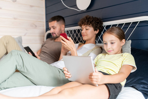 Милая маленькая девочка в повседневной одежде, используя планшет, сидя на кровати рядом со своими родителями со смартфонами, просматривающими сообщения в социальных сетях - Фото, изображение