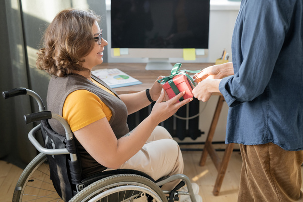 Щаслива молода бізнес-леді в інвалідному візку, приймаючи упаковану подарункову коробку з подарунком на Різдво або день народження з рук її колеги в офісі
 - Фото, зображення