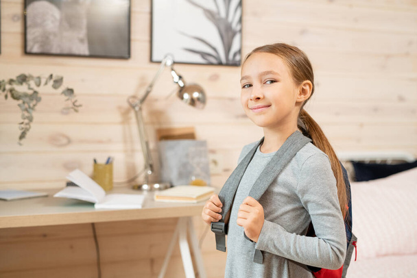 Contemporary happy schoolgirl with backpack standing in front of camera against her bed and table with books by wooden wall with two pictures - Photo, Image