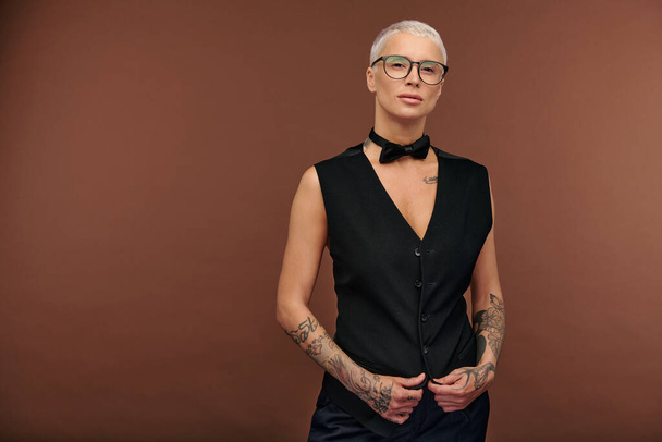 Serene masculine elegant blond female in black waistcoat and bowtie standing against brown background in isolation in front of camera - Photo, Image