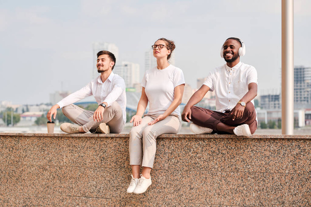 Groupe de jeunes cols blancs sereins et détendus en tenue décontractée assis sur un mur de marbre carrelé au bord de la rivière et méditant à la pause - Photo, image