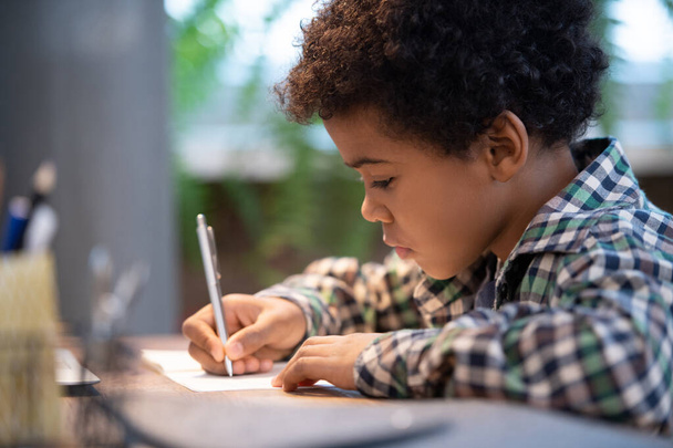 Widok z boku cute little African ucznia w casualwear dokonywania notatek w copybook siedząc przy stole i wykonywania zadań domowych - Zdjęcie, obraz