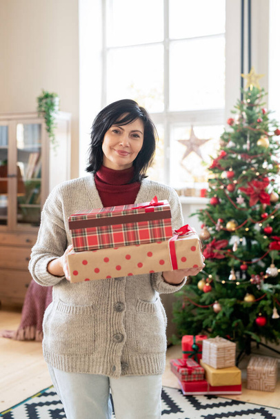 Szczęśliwy Dojrzałe kobieta w casualwear gospodarstwa pudełka z prezentami dla jej rodziny, stojąc przeciwko urządzone choinki w domu - Zdjęcie, obraz