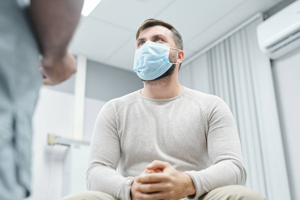 Симпатичный пациент-мужчина в сером свитере и защитной маске смотрит на врача и слушает объяснение его медицинских результатов в клиниках - Фото, изображение