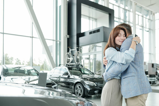 Felice giovane donna bruna in casualwear intelligente che abbraccia il marito dopo l'acquisto di auto nuove nel centro auto lussuoso contemporaneo - Foto, immagini