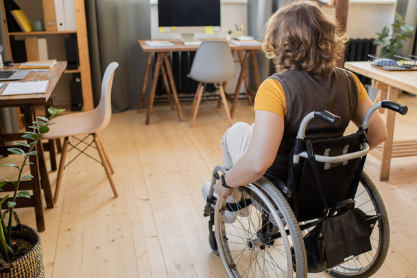 Rückansicht der jungen brünetten behinderten Frau, die im Rollstuhl vor der Kamera sitzt, während sie sich auf einen der Tische mit Computermonitor zubewegt - Foto, Bild