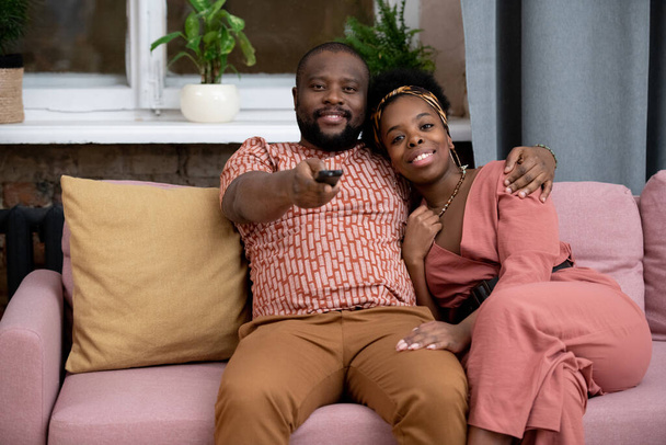 Feliz joven afectuoso marido africano y esposa sentado en el sofá contra la ventana en la sala de estar y viendo el programa de televisión o película - Foto, imagen