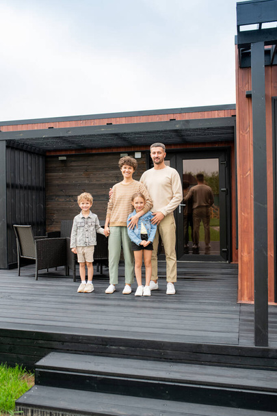 Szczęśliwa współczesna rodzina młodych rodziców i ich dwoje uroczych małych dzieci stojących na patio nowego domu na tle czarnych foteli - Zdjęcie, obraz