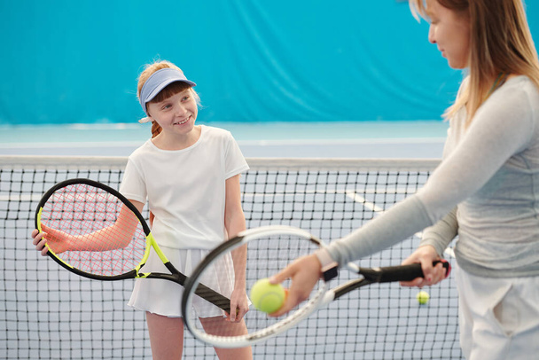 Hymyilevä aktiivinen tyttö tennismailalla katsomalla naisten kouluttaja selittää hänelle, miten käyttää sitä pelin aikana, jos haluat olla menestyksekäs pelaaja - Valokuva, kuva