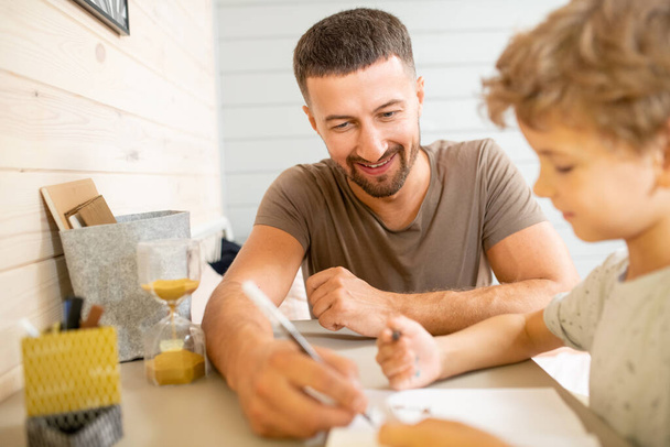 Junger glücklicher Vater mit Buntstift hilft seinem kleinen Sohn beim Zeichnen, während er auf dem Bett am Tisch sitzt und beide neue kreative Ideen diskutieren - Foto, Bild