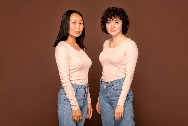 Δύο νεαρές διαπολιτισμικές γυναίκες με λευκά πουλόβερ και μπλε τζιν να στέκονται η μία δίπλα στην άλλη με καφέ φόντο και να σε κοιτάζουν - Φωτογραφία, εικόνα