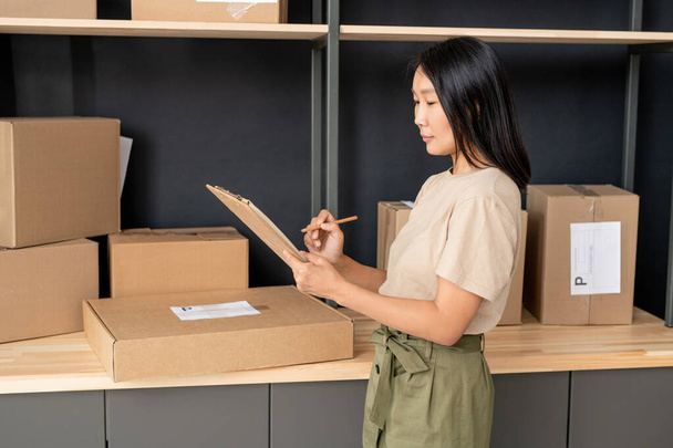 Young Asian female with pen and clipboard checking orders of clients of online shop while working in store-room against shelf with boxes - Photo, Image