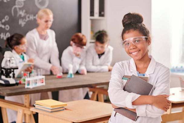 Menina da escola secundária mista feliz em whitecoat olhando para você com sorriso de dente contra colegas de classe e professor durante o trabalho de laboratório - Foto, Imagem