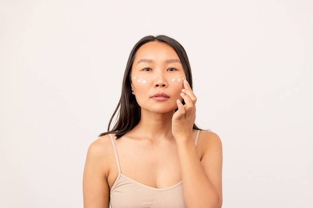 Chica coreana o china con el pelo largo oscuro que tiene máscara debajo de los ojos mientras cuida de su piel delante de la cámara sobre fondo blanco - Foto, imagen
