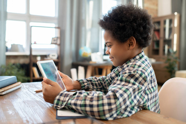 Seitenansicht eines afrikanischen Grundschülers mit Touchpad, der im Wohnzimmer am Tisch sitzt und Online-Unterricht verfolgt, um Informationen für die Schule zu erhalten - Foto, Bild