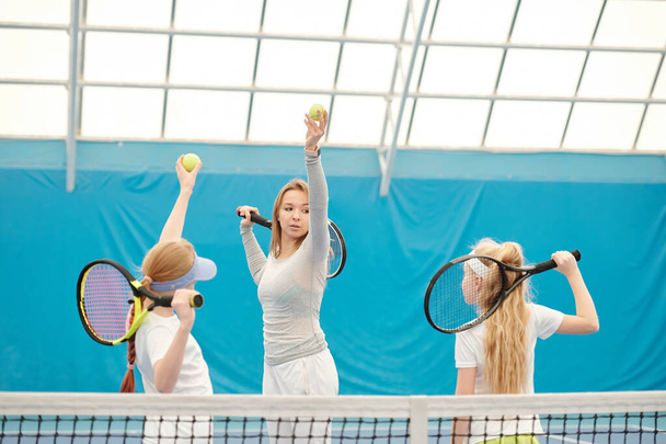 Молодой тренер по теннису в спортивной одежде держит ракетку за шеей и поднимает руку с мячом, консультируясь с двумя симпатичными девушками на стадионе - Фото, изображение