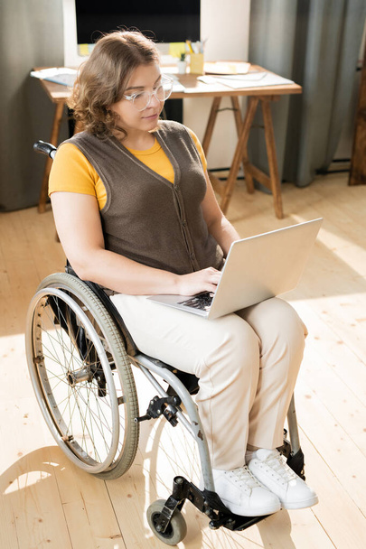 Pretty young disable woman in casualwear typing on laptop keypad while surfing in the net or during remote work in the center of domestic room - Photo, Image