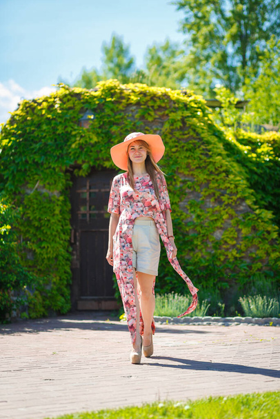 A charming girl in a light summer sundress and a pareo hat is walking in a green park. Enjoys warm sunny summer days. - Φωτογραφία, εικόνα