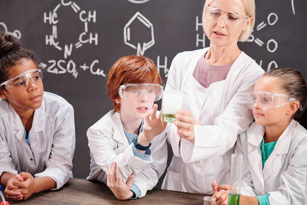 Giovane insegnante di chimica mostrando intelligenti alunni miscela di sostanze chimiche a lezione mentre curioso scolaro toccare becco - Foto, immagini