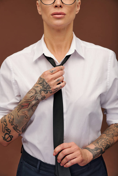 Kortárs középkorú üzletasszony tetoválással a karján, fekete nyakkendőt visel, miközben elszigetelten áll a kamera előtt. - Fotó, kép