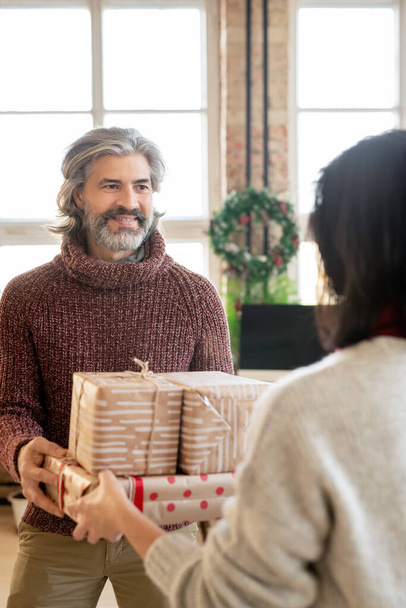 Щасливий зрілий чоловік бере подарункові коробки з рук своєї дружини, дивлячись на неї проти різдвяного вінка на великому вікні в домашньому середовищі
 - Фото, зображення