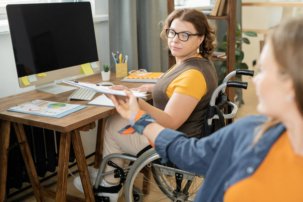 Młoda poważna bizneswoman na wózku inwalidzkim biorąc schowek z dokumentem finansowym z rąk kolegi podczas pracy z danymi w biurze - Zdjęcie, obraz