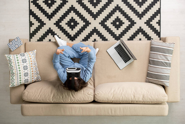 Vista general de la joven morena en pijama azul y auriculares vr sentado en un sofá suave con las piernas cruzadas y conmovedora pantalla virtual - Foto, imagen