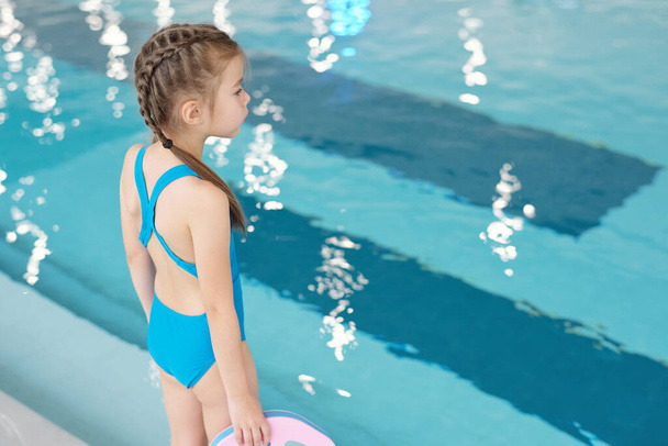 Милая маленькая девочка в синих купальниках смотрит на прозрачную воду в бассейне, стоя перед камерой напротив бассейна в спортивном центре - Фото, изображение