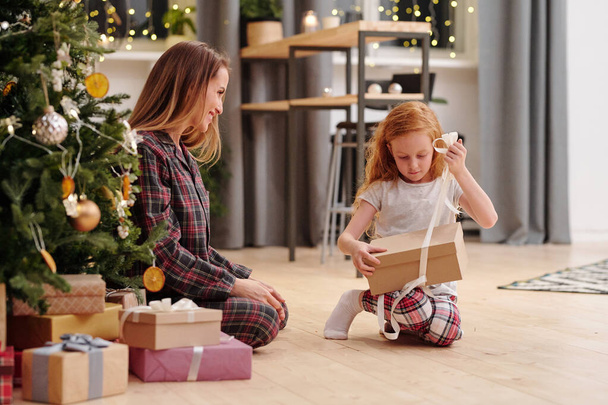 Cute little girl untying ribbon on packed giftbox while going to take out xmas present from her mother sitting on the floor by decorated firtree - Photo, Image