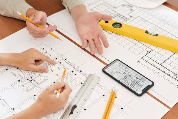 Handen van twee jonge ingenieurs met potloden over schetsen van nieuwbouw tijdens de bespreking van de details en creatieve ideeën - Foto, afbeelding