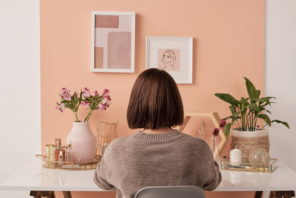Вид ззаду брюнетка дизайнерка інтер'єру сидить за столом перед рожевою стіною з двома фотографіями в рамах і виконує свою роботу
 - Фото, зображення