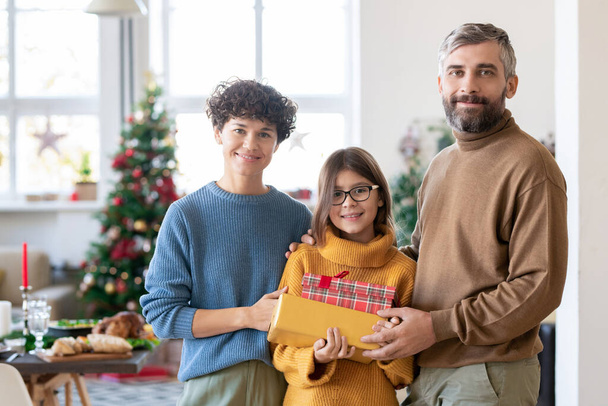 Веселая молодая пара и их дочь-подросток держат в руках подарочные коробки с подарками, стоя напротив украшенной елки в гостиной - Фото, изображение