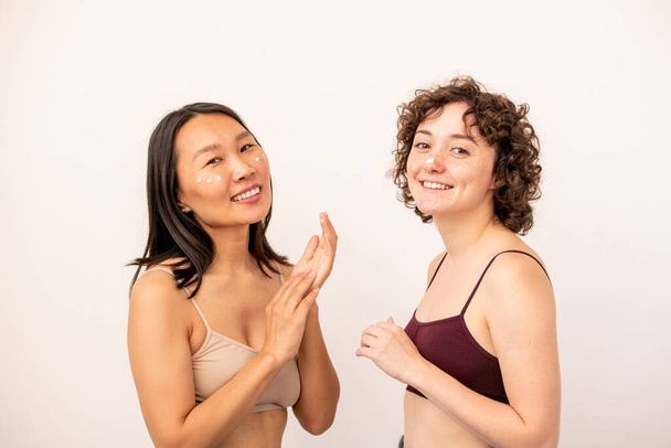 Дві молоді веселі міжкультурні жінки на сонячних батареях наносять омолоджуючий крем на неприємну ділянку, стоячи перед камерою
 - Фото, зображення