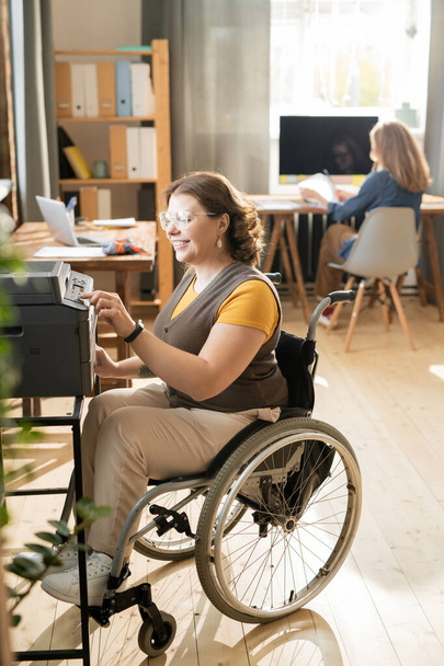 Gelukkig jong uitschakelen vrouw in rolstoel zitten door xerox machine en druk op de startknop, terwijl het maken van kopieën van documenten in het kantoor - Foto, afbeelding