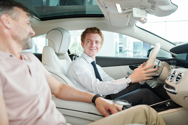 Happy young sales manager in formalwear sitting in new car and holding by steer during test drive with mature male client sitting next to him - Fotografie, Obrázek
