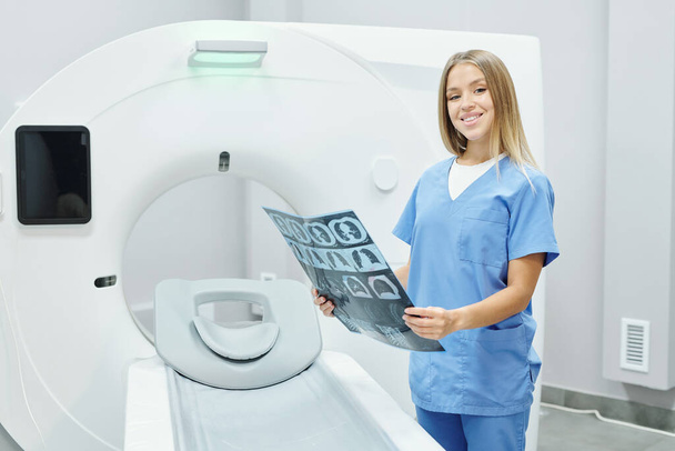 Ευτυχισμένη νεαρή ξανθιά ακτινολογός με μπλε στολή αναλύοντας την ακτινογραφία της κεφαλής του ασθενούς ενάντια στο υπερηχητικό μηχάνημα στο ιατρικό γραφείο - Φωτογραφία, εικόνα