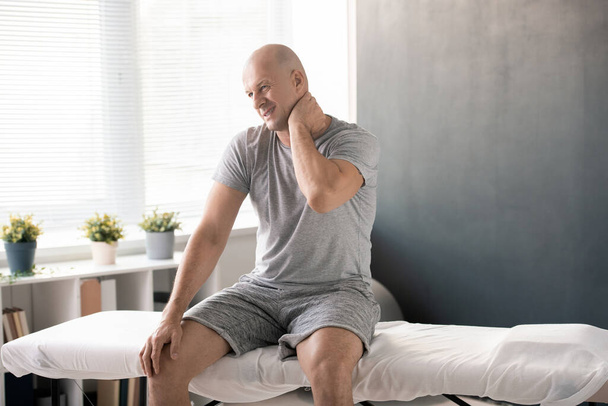 Plešatý muž dotýká bolestivý krk na levé straně a snaží se masírovat před fyzickým cvičením, zatímco sedí na gauči a čeká na lékaře - Fotografie, Obrázek