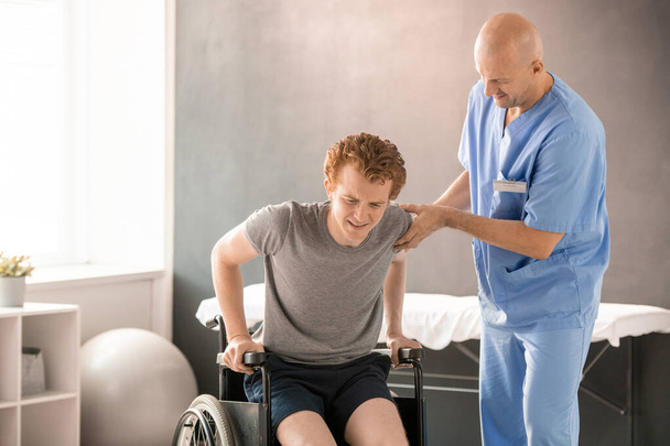 Dojrzały klinicysta w mundurze pomaga młodzieńcowi w bólu usiąść na wózku inwalidzkim po treningu rehabilitacyjnym wspierając jednocześnie jego ramię i łokieć - Zdjęcie, obraz