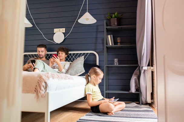 Serieus klein meisje met touchpad kijken naar online video terwijl zitten op de vloer door bed waar haar ouders scrollen door nieuws in smartphones - Foto, afbeelding