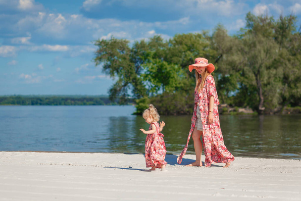 A charming girl in a light summer sundress walks on the sandy beach with her little daughter. Enjoys warm sunny summer days. - Фото, зображення