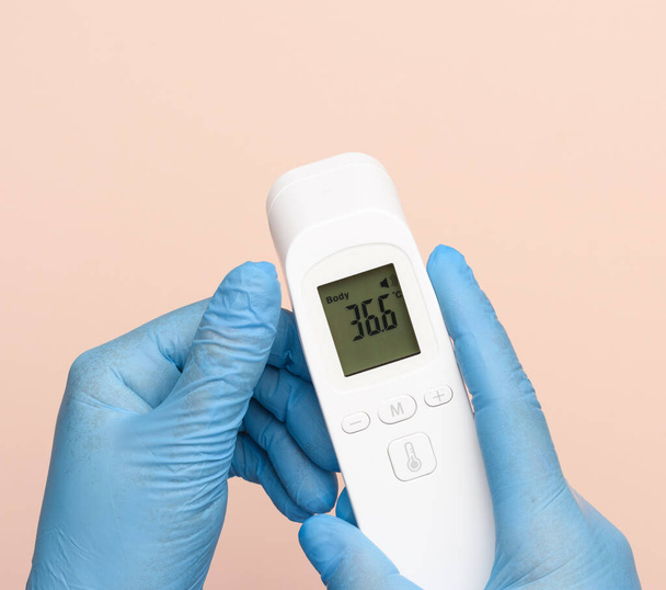 Hand in blauen Latex-Handschuhen hält ein elektronisches Thermometer zur Temperaturmessung, berührungsloses Gerät, Anzeige zeigt Temperatur 36,6 Grad  - Foto, Bild