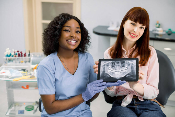 Shot of professional female african dentist and caucasian woman patient sitting in dentistry chair, showing digital tablet with patient oral scan, jaws and teeth x-ray, to camera and smiling - Photo, image