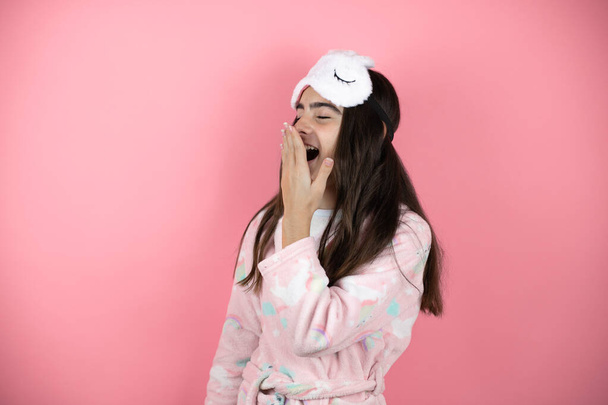 Ładna dziewczyna w piżamie i masce na różowym tle znudzona ziewaniem zmęczona przykrywaniem ust ręką. Niepokój i senność. - Zdjęcie, obraz